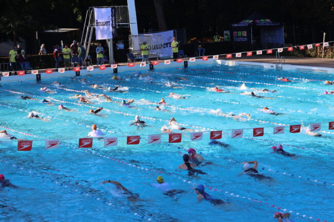 Zwembad Buitenzwembad met zwemmers tussen de lijnen en met half schaduw en half in de zon. Speedo vlaggetjes boven het water. Spandoek met sponsor Brouwers op de achtergrond