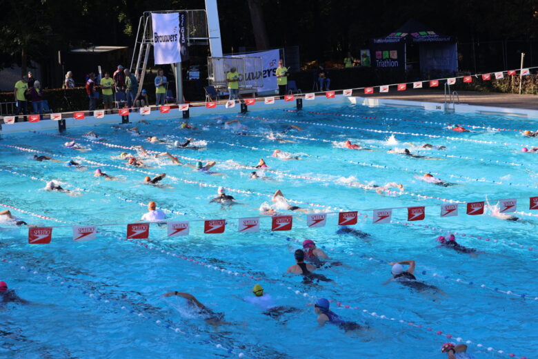 Buitenzwembad met zwemmers tussen de lijnen en met half schaduw en half in de zon. Speedo vlaggetjes boven het water. Spandoek met sponsor Brouwers op de achtergrond